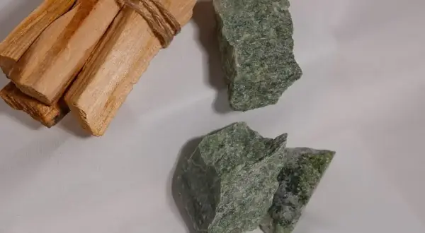 Surowe bryłki zielonego jadeitu, kamienia o licznych właściwościach, leżą obok wiązki drewna palo santo.
