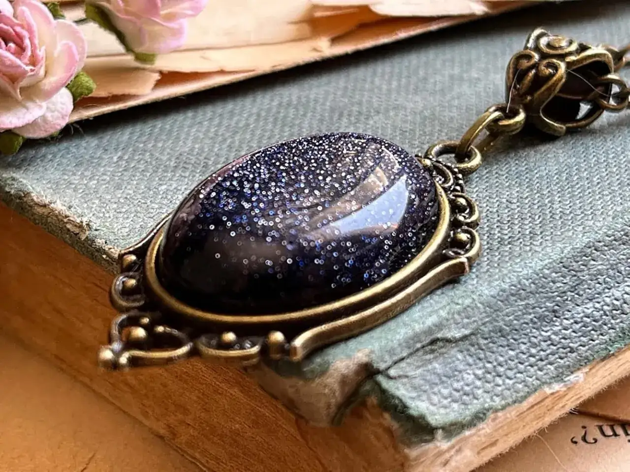 Amulet z granatowym kamieniem, iskrzącym jak noc kairu. Delikatne róże i stara księga tworzą magiczną scenerię.